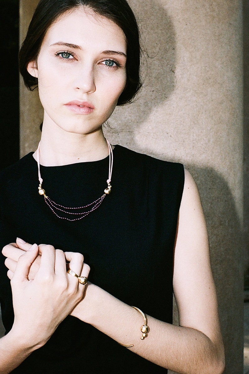 May include: A woman wearing a black sleeveless dress with a gold necklace and bracelet. The necklace has a pink cord and small gold beads. The bracelet is a simple gold band with two small gold balls.