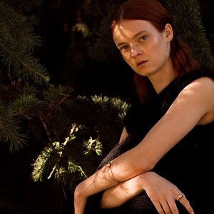 May include: A woman with red hair wearing a black sleeveless top and gold jewelry. She is sitting in front of a pine tree.