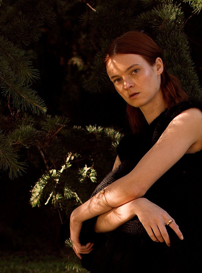 May include: A woman with red hair wearing a black sleeveless top and gold jewelry. She is sitting in front of a pine tree.