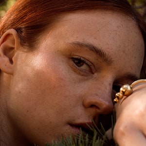 May include: Close-up of a woman's face with freckles, red hair, and a gold ring on her finger. Her eyes are closed and she is looking down. The background is blurred and out of focus.