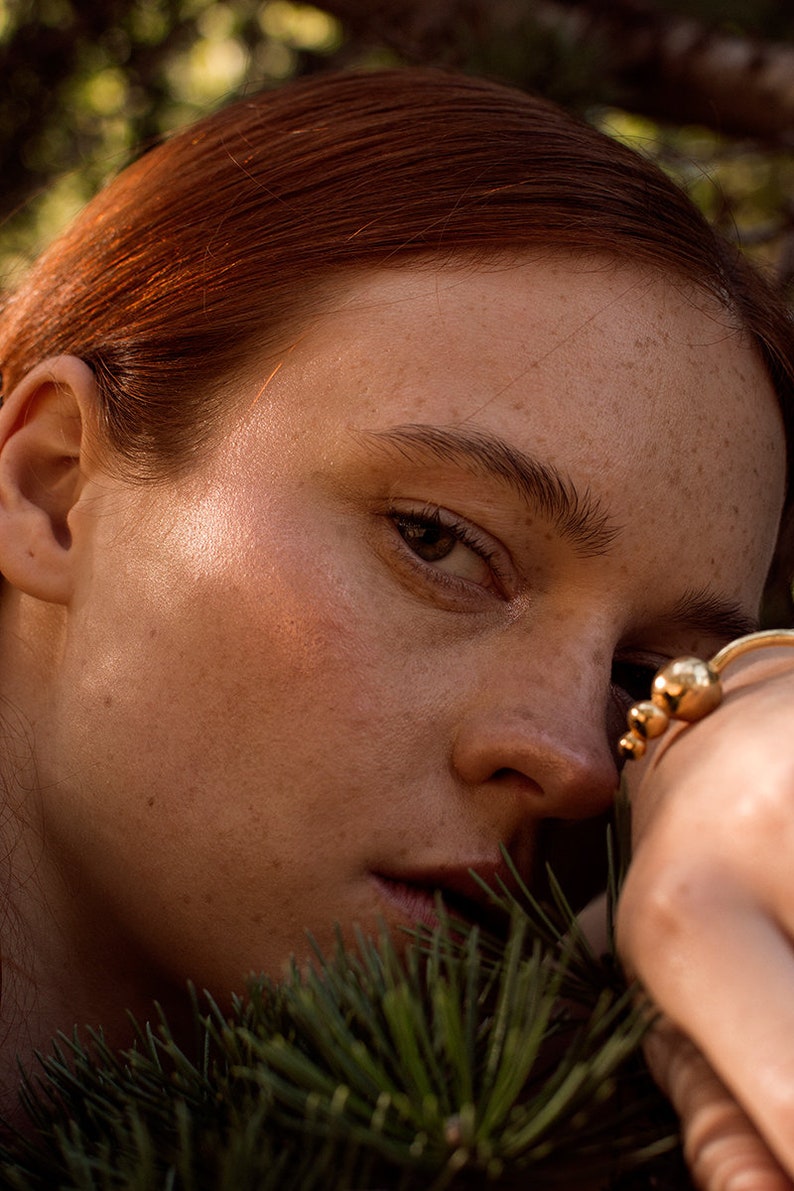 May include: Close-up of a woman's face with freckles, red hair, and a gold ring on her finger. Her eyes are closed and she is looking down. The background is blurred and out of focus.