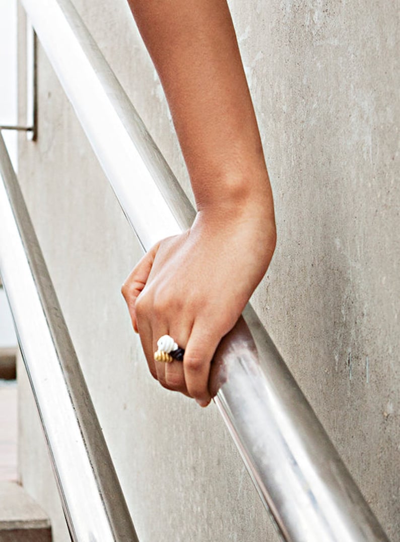 May include: A close-up of a hand wearing a gold ring with white and black accents. The ring is made of multiple small, round beads.