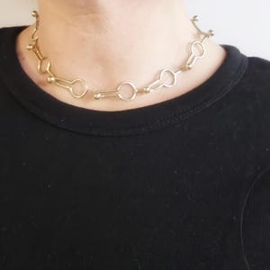 May include: A gold-colored necklace with a unique design. The necklace features a series of connected keyhole-shaped links, interspersed with small, round beads. The necklace is worn against a black shirt.