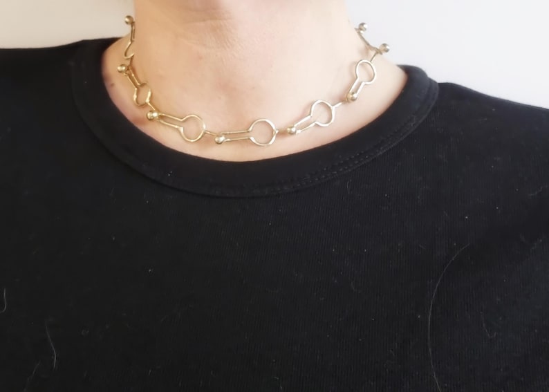 May include: A gold-colored necklace with a unique design. The necklace features a series of connected keyhole-shaped links, interspersed with small, round beads. The necklace is worn against a black shirt.
