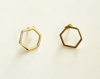 May include: A pair of gold-colored, open-frame hexagon stud earrings. The earrings are geometric in design, with a simple, modern aesthetic. The earrings are displayed on a white surface.