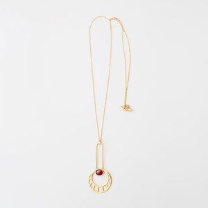 May include: A gold necklace with a geometric pendant featuring a red stone. The pendant is a circle with a vertical bar extending from the top. The circle has a cutout design and the stone is set in the center.