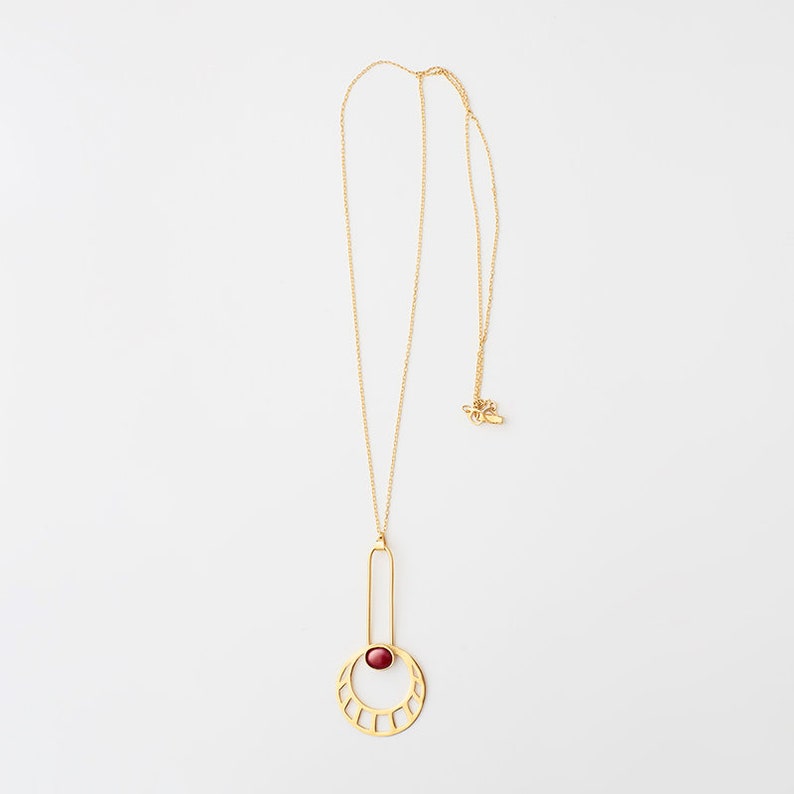 May include: A gold necklace with a geometric pendant featuring a red stone. The pendant is a circle with a vertical bar extending from the top. The circle has a cutout design and the stone is set in the center.