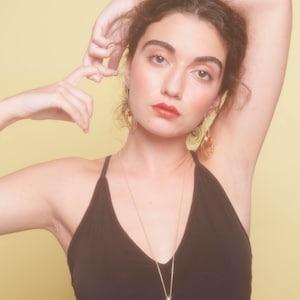 May include: A woman wearing a black tank top with a gold necklace featuring a red gemstone and a geometric design. She is wearing gold earrings with red gemstones.