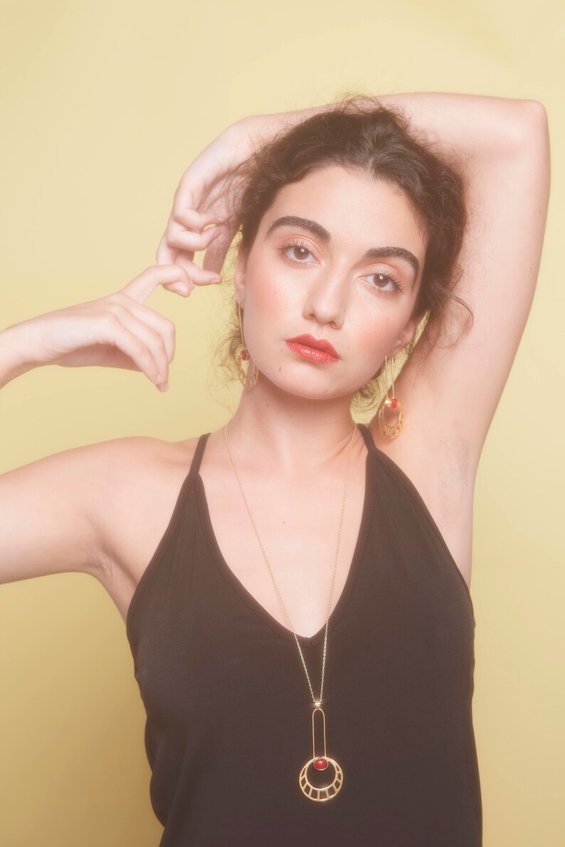 May include: A woman wearing a black tank top with a gold necklace featuring a red gemstone and a geometric design. She is wearing gold earrings with red gemstones.