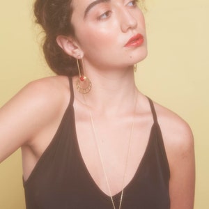 May include: A woman wearing a black tank top with a gold necklace and earrings featuring a red gemstone. The necklace and earrings have a geometric design.