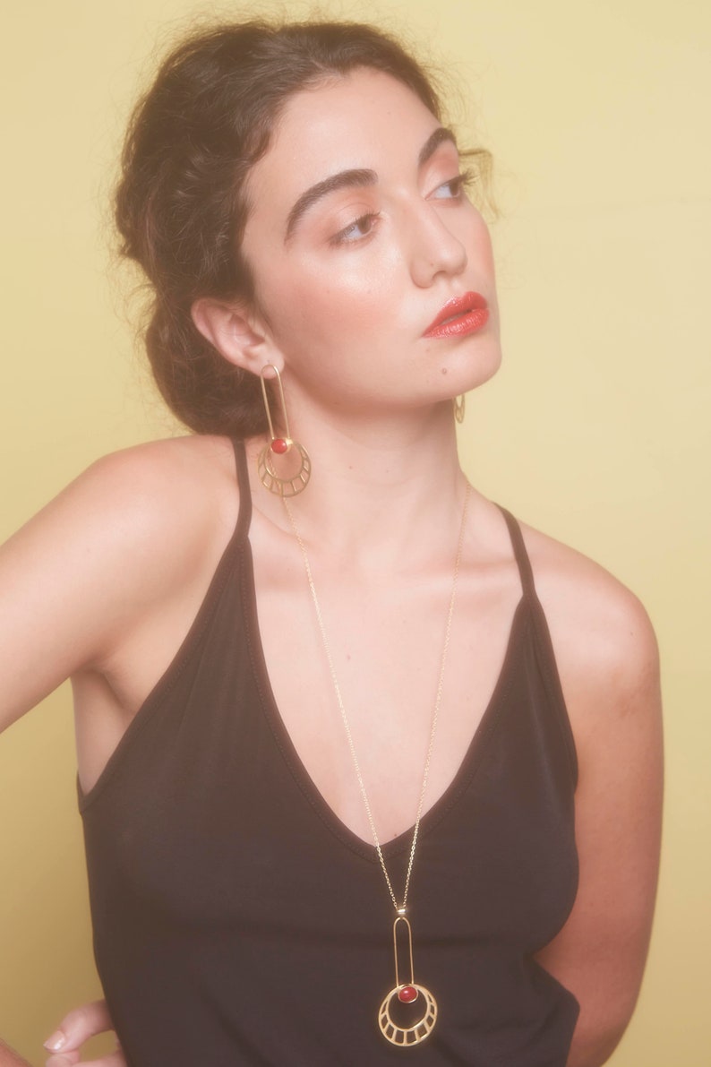 May include: A woman wearing a black tank top with a gold necklace and earrings featuring a red gemstone. The necklace and earrings have a geometric design.