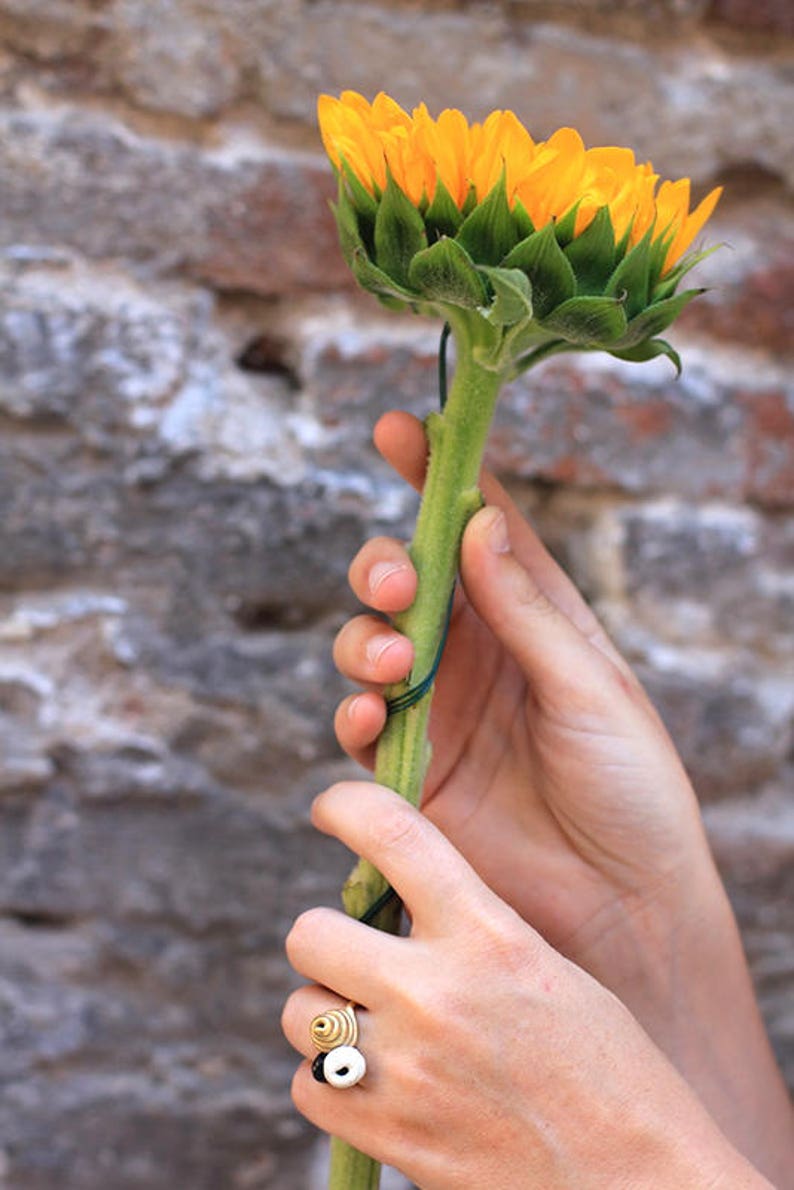 May include: A hand wearing a gold ring with black and white accents holds a sunflower with yellow petals and green leaves.