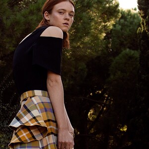 May include: A woman wearing a black off-the-shoulder top and a plaid skirt with ruffles. She is standing in a forest setting.