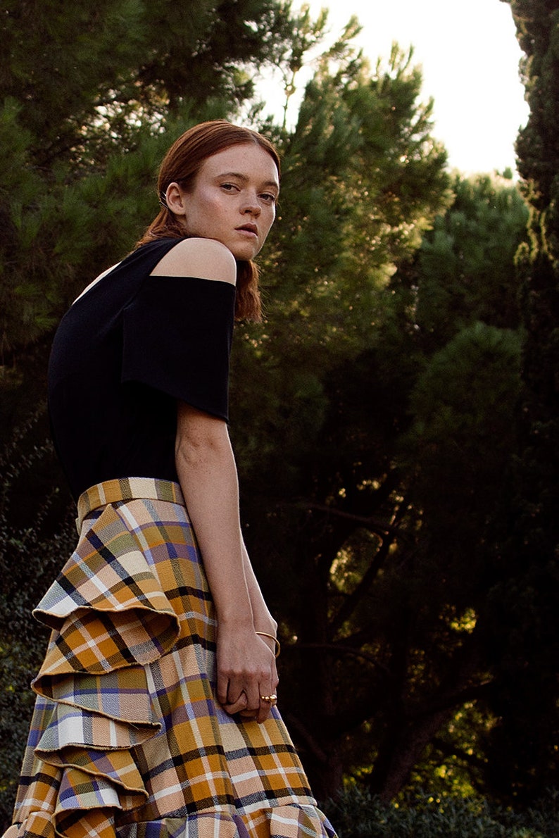 May include: A woman wearing a black off-the-shoulder top and a plaid skirt with ruffles. She is standing in a forest setting.