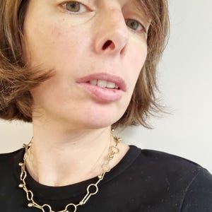 May include: A gold-colored necklace with a unique design featuring a series of connected circles and small, round accents. The necklace is worn against a black top, creating a contrast that highlights its intricate details. The jewelry is a statement piece.