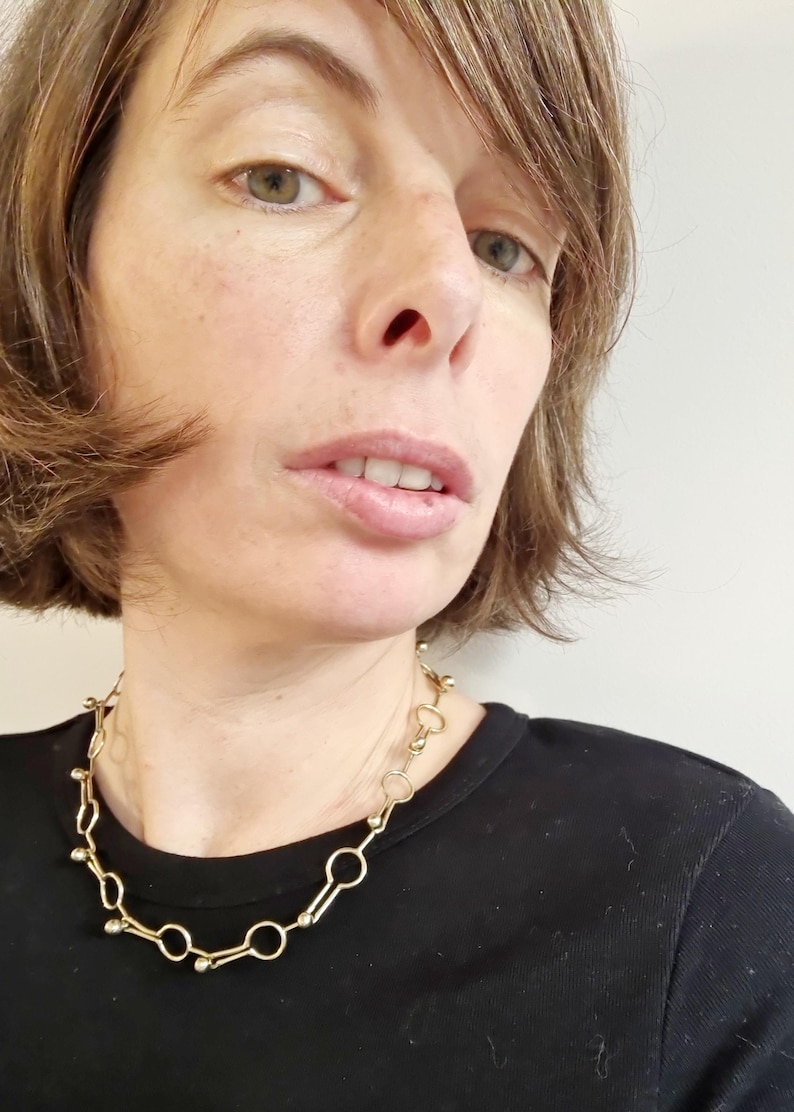 May include: A gold-colored necklace with a unique design featuring a series of connected circles and small, round accents. The necklace is worn against a black top, creating a contrast that highlights its intricate details. The jewelry is a statement piece.