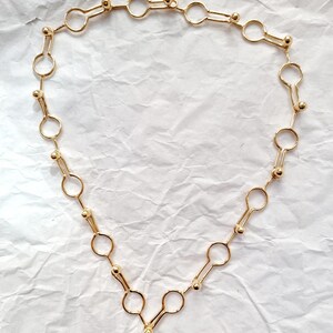 May include: A gold-colored necklace with a unique design. The necklace features a series of connected shapes, including circles and elongated elements with small ball accents. The jewelry is displayed on a white, textured surface.