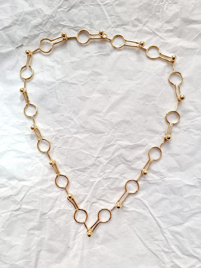 May include: A gold-colored necklace with a unique design. The necklace features a series of connected shapes, including circles and elongated elements with small ball accents. The jewelry is displayed on a white, textured surface.