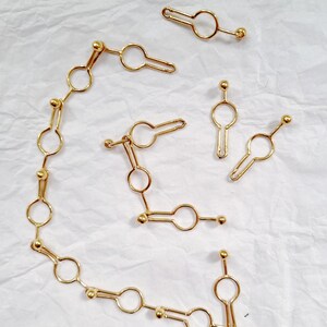 May include: Gold-colored jewelry pieces arranged on a white surface. The pieces include a chain of connected circles and key-shaped elements, along with several individual key-shaped charms. The jewelry has a shiny finish.
