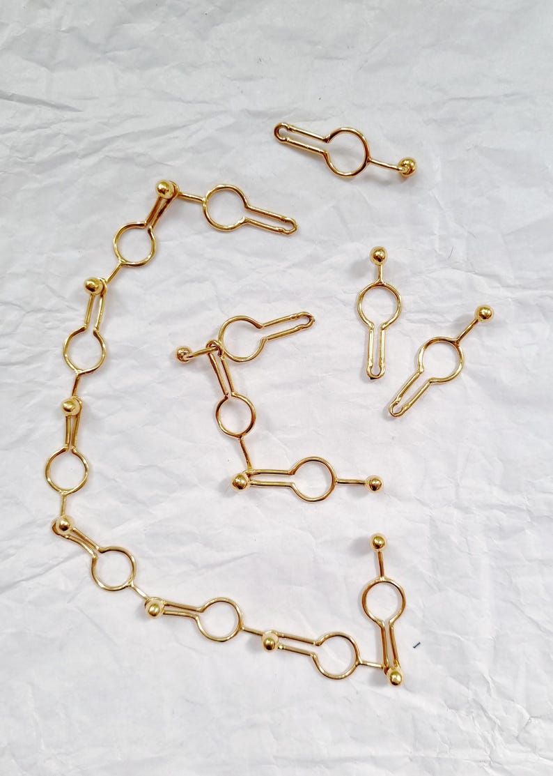 May include: Gold-colored jewelry pieces arranged on a white surface. The pieces include a chain of connected circles and key-shaped elements, along with several individual key-shaped charms. The jewelry has a shiny finish.