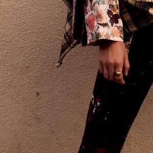 May include: A close-up of a person's hand wearing a gold ring. The hand is wearing black pants with a white and floral print. The person is wearing a brown and black patterned jacket.