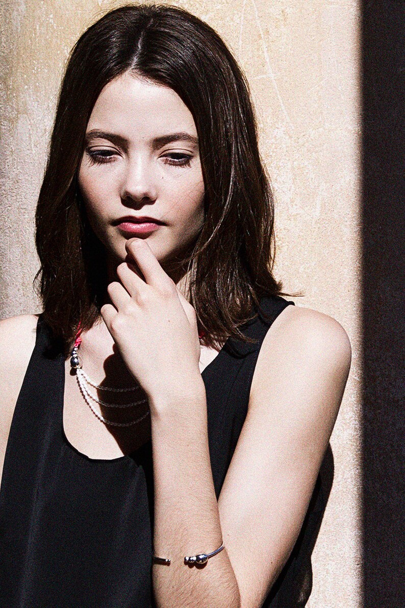 May include: A woman with long brown hair is wearing a black tank top and a silver bracelet with three round beads. She is wearing a layered necklace with a silver charm.