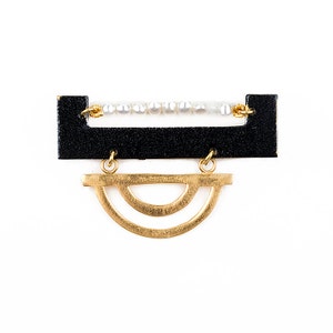 May include: A black and gold geometric brooch with a row of white pearls. The brooch features a rectangular shape with a semi-circle design hanging from the bottom.
