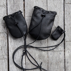 May include: Three black leather pouches with drawstrings and a metal clasp. The pouches are on a wooden surface.