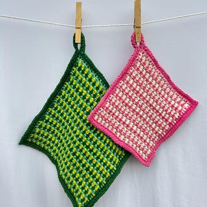 May include: Two handmade crochet potholders hanging from a clothesline. One is green with yellow accents and a dark green border. The other is pink with white accents and a pink border. Both are square with a loop for hanging.