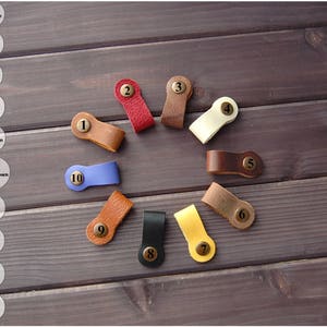 K&ouml;nnte beinhalten: Zehn Leder-Kabelbinder mit nummerierten Messing-Etiketten. Die Kabelbinder sind in verschiedenen Farben, darunter Braun, Beige, Rot, Blau und Gelb.