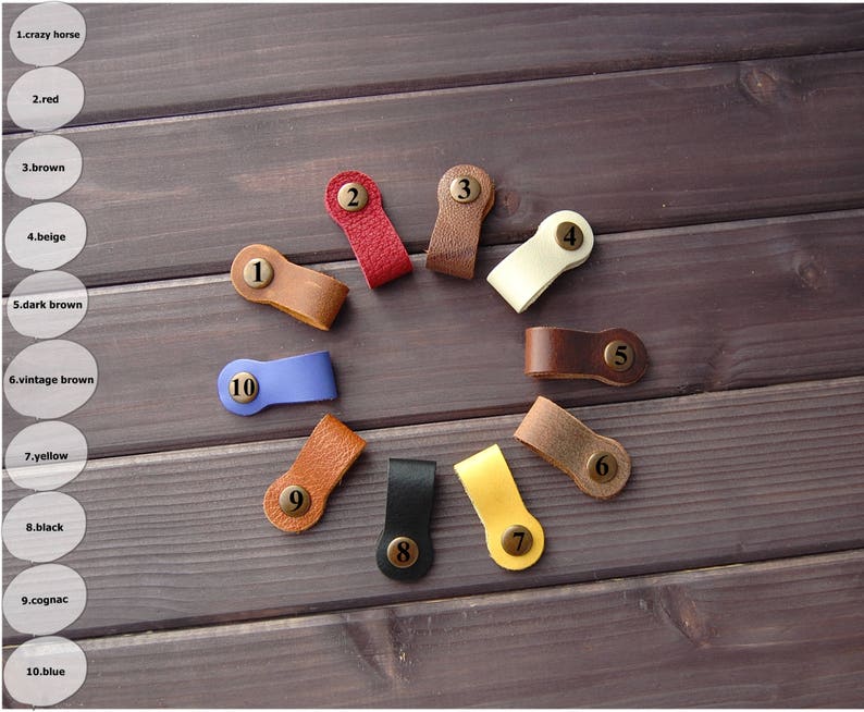 K&ouml;nnte beinhalten: Zehn Leder-Kabelbinder mit nummerierten Messing-Etiketten. Die Kabelbinder sind in verschiedenen Farben, darunter Braun, Beige, Rot, Blau und Gelb.