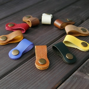 K&ouml;nnte beinhalten: Sortiment an Leder-Kabelorganisatoren in verschiedenen Farben, darunter Blau, Gelb, Braun, Rot und Schwarz. Jeder Organizer hat einen Metallknopf zum Sichern von Kabeln.
