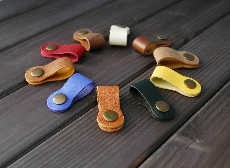 K&ouml;nnte beinhalten: Sortiment an Leder-Kabelorganisatoren in verschiedenen Farben, darunter Blau, Gelb, Braun, Rot und Schwarz. Jeder Organizer hat einen Metallknopf zum Sichern von Kabeln.