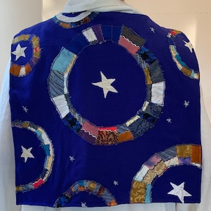 Könnte beinhalten: Eine blaue Stoffweste mit einem Patchwork-Muster aus Sternen und Kreisen. Die Kreise bestehen aus verschiedenen Stoffen und sind kreisförmig angeordnet. Die Sterne sind weiß und auf den Stoff gestickt.