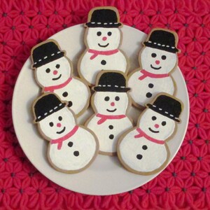 Puede incluir: Seis galletas de az&uacute;car con forma de mu&ntilde;eco de nieve decoradas con glaseado blanco y negro. Las galletas est&aacute;n dispuestas en un plato blanco con un fondo de crochet rojo.