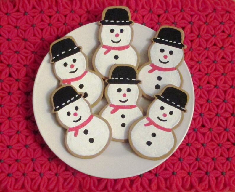 Puede incluir: Seis galletas de az&uacute;car con forma de mu&ntilde;eco de nieve decoradas con glaseado blanco y negro. Las galletas est&aacute;n dispuestas en un plato blanco con un fondo de crochet rojo.