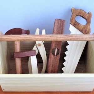 May include: A wooden toy toolbox with a handle and five wooden tools inside: a hammer, a saw, a level, a pair of pliers, and a clamp.