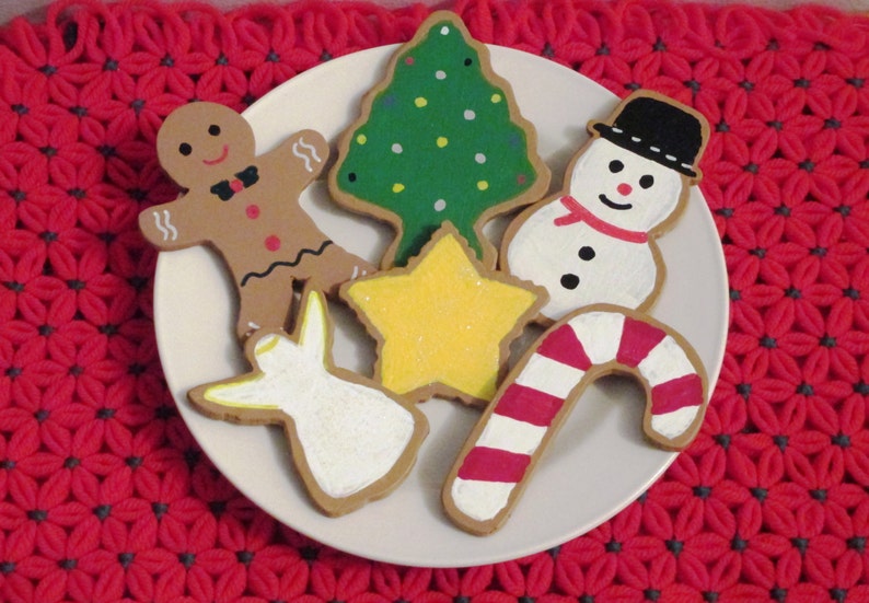 Puede incluir: Un conjunto de seis galletas de az&uacute;car con tem&aacute;tica navide&ntilde;a decoradas con glaseado. Las galletas tienen forma de hombre de jengibre, &aacute;rbol de Navidad, mu&ntilde;eco de nieve, bast&oacute;n de caramelo, estrella y &aacute;ngel. Las galletas est&aacute;n en un plato blanco.