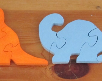 May include: Four wooden dinosaur puzzle pieces in different colors: green, orange, blue, and yellow. Each piece has a unique shape and is designed to fit together.