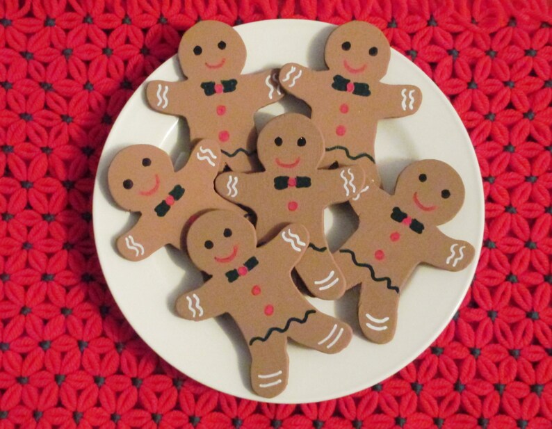Puede incluir: Seis galletas de jengibre en forma de hombre en un plato blanco. Las galletas est&aacute;n decoradas con ojos de glaseado negro, sonrisas de glaseado rojo y corbatas de lazo de glaseado negro. El plato est&aacute; sobre una superficie de crochet roja y gris.