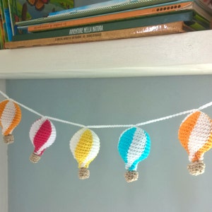 May include: A garland of crocheted hot air balloons in orange, white, red, yellow, and blue hangs from a white shelf. The balloons are strung together with white string.