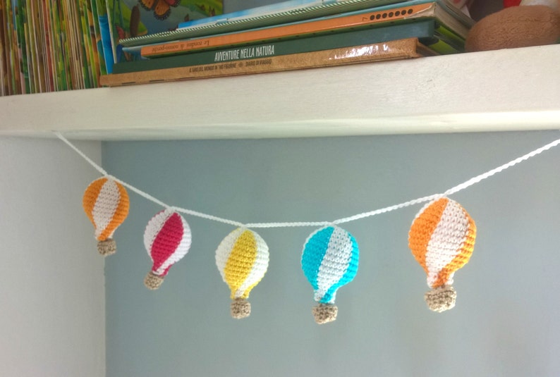 May include: A garland of crocheted hot air balloons in orange, white, red, yellow, and blue hangs from a white shelf. The balloons are strung together with white string.