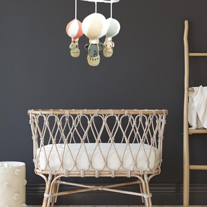 May include: A white wicker crib with a white mattress and a mobile hanging above it. The mobile has three crocheted hot air balloons with animal faces and a white base.