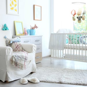 May include: A white nursery with a white crib, a white armchair with a plaid blanket, a dresser with drawers, and a mobile hanging above the crib. The mobile has hot air balloons and animal figures.