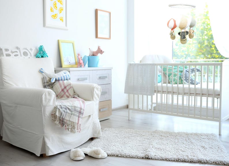 May include: A white nursery with a white crib, a white armchair with a plaid blanket, a dresser with drawers, and a mobile hanging above the crib. The mobile has hot air balloons and animal figures.