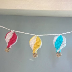 May include: A garland of five crocheted hot air balloons in orange, pink, yellow, and blue with white stripes. The balloons are strung together with white yarn.