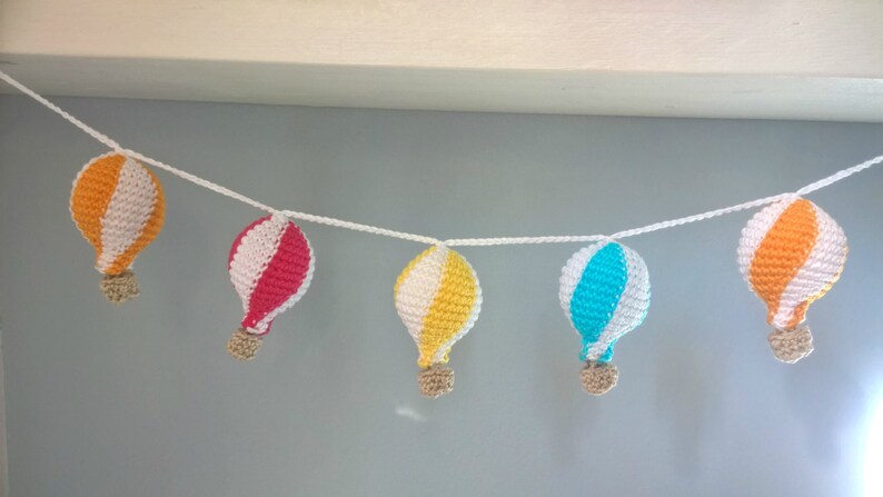 May include: A garland of five crocheted hot air balloons in orange, pink, yellow, and blue with white stripes. The balloons are strung together with white yarn.