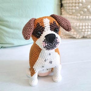 Könnte beinhalten: Ein gehäkeltes Stofftier in Form eines Hundes mit braunem, weißem und grauem Fell. Der Hund sitzt mit leicht geneigtem Kopf und blickt direkt in die Kamera.