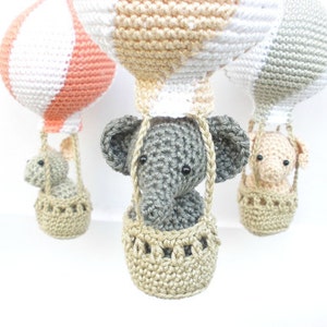 May include: Three crocheted hot air balloons with animal passengers. The balloons are in shades of peach, white, and gray. The animals are a gray elephant, a gray bunny, and a peach-colored pig. Each animal is in a crocheted basket.