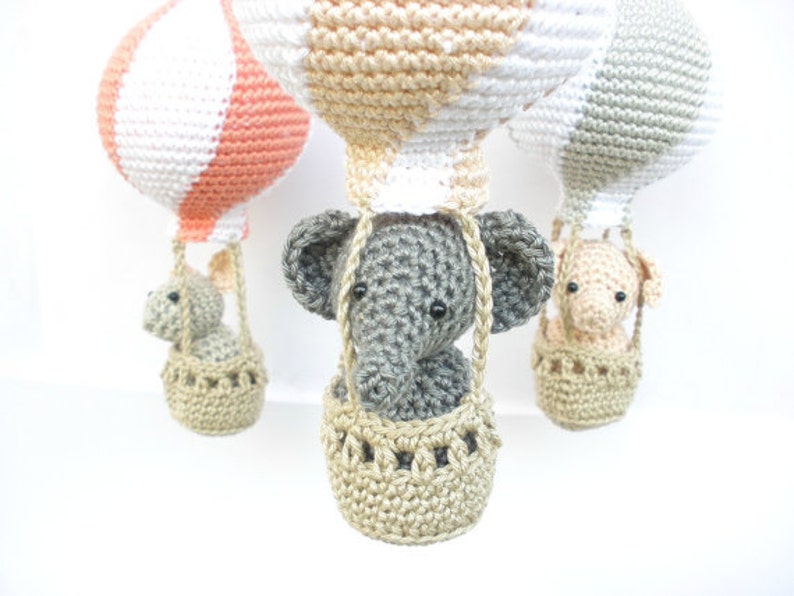 May include: Three crocheted hot air balloons with animal passengers. The balloons are in shades of peach, white, and gray. The animals are a gray elephant, a gray bunny, and a peach-colored pig. Each animal is in a crocheted basket.